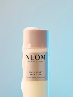 NEOM Real Luxury bath foam with lavender, jasmine and sandalwood surrounded by bubbles on blue background.
