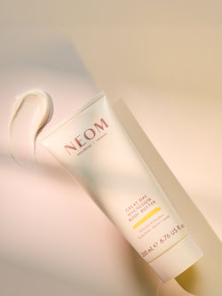 NEOM Great Day magnesium body butter tube with creamy texture swatch on warm pastel background.