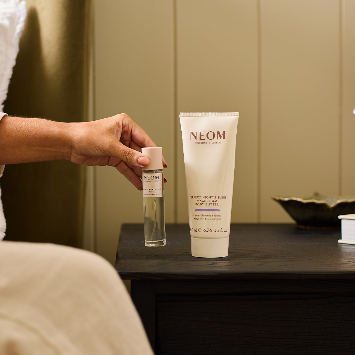 A person putting NEOM perfect nights sleep mist on a bedside table with the magnesium body butter next to it