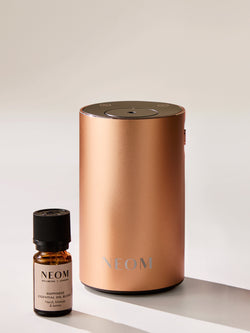 NEOM wellbeing pod diffuser in rose gold with Happiness essential oil blend on minimal studio background.