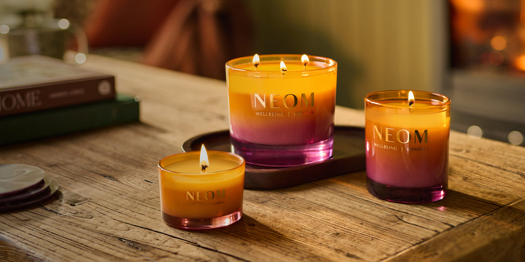 Three NEOM Cosy Nights candles on a wooden surface with a blurred background