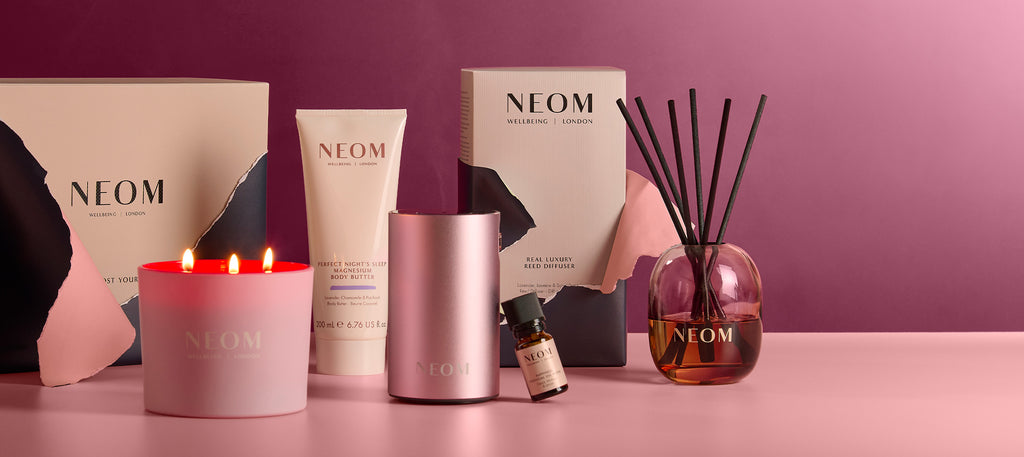 NEOM wellbeing gifts featuring It's All Rosy candle, magnesium body butter, reed diffuser, essential oil, and Pink Wellbeing Pod Mini+ on pink background.