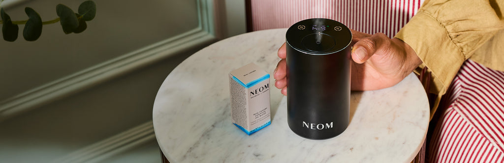 Person holding a black NEOM Pod Mini+ on a marble table with a box next to it.