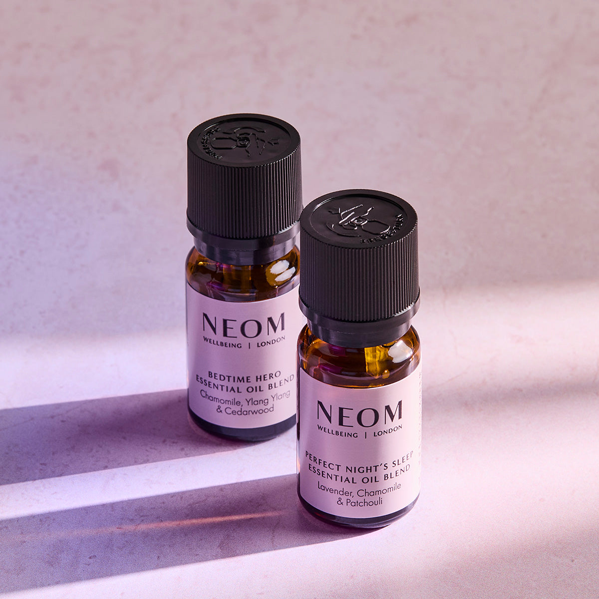 Two NEOM essential oil blend bottles, Bedtime Hero and Perfect Night’s Sleep, standing on a softly lit surface.
