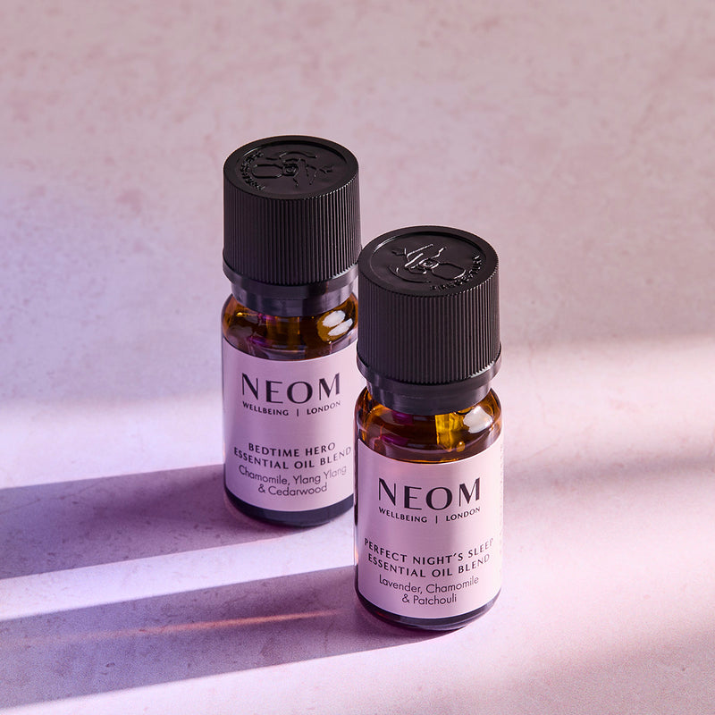 Two NEOM essential oil blend bottles, Bedtime Hero and Perfect Night’s Sleep, standing on a softly lit surface.