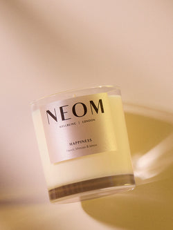 NEOM Happiness scented candle with neroli, mimosa and lemon in glass jar on warm neutral background.