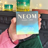 Hand holding NEOM Ceramic Circle Natural Diffuser box with green candle and festive lights in background.