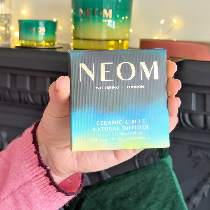 Hand holding NEOM Ceramic Circle Natural Diffuser box with green candle and festive lights in background.