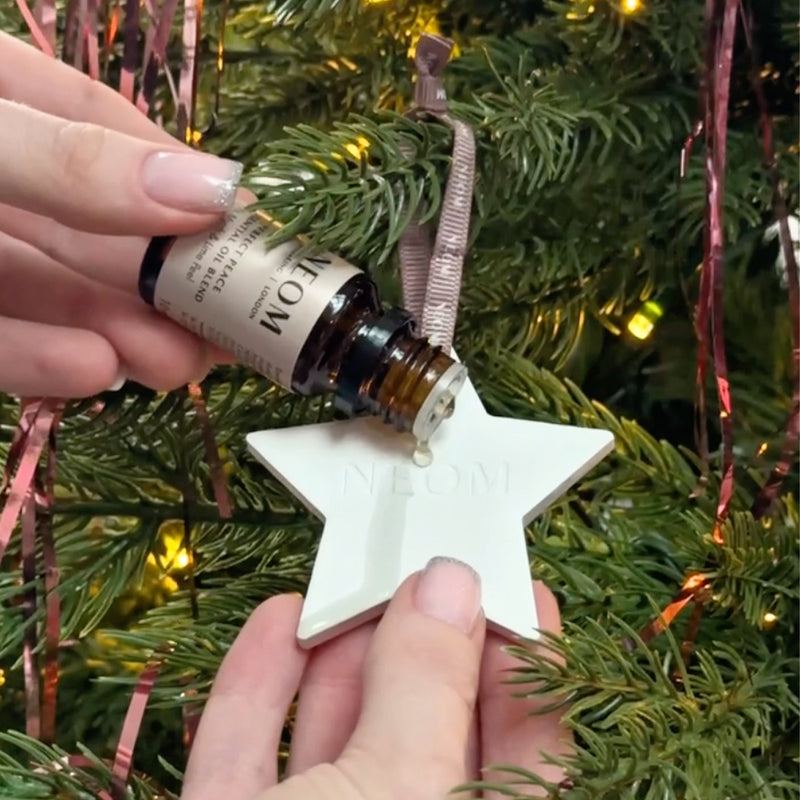 Person adding NEOM Essential Oil Blend to ceramic star ornament on Christmas tree with pink ribbon.
