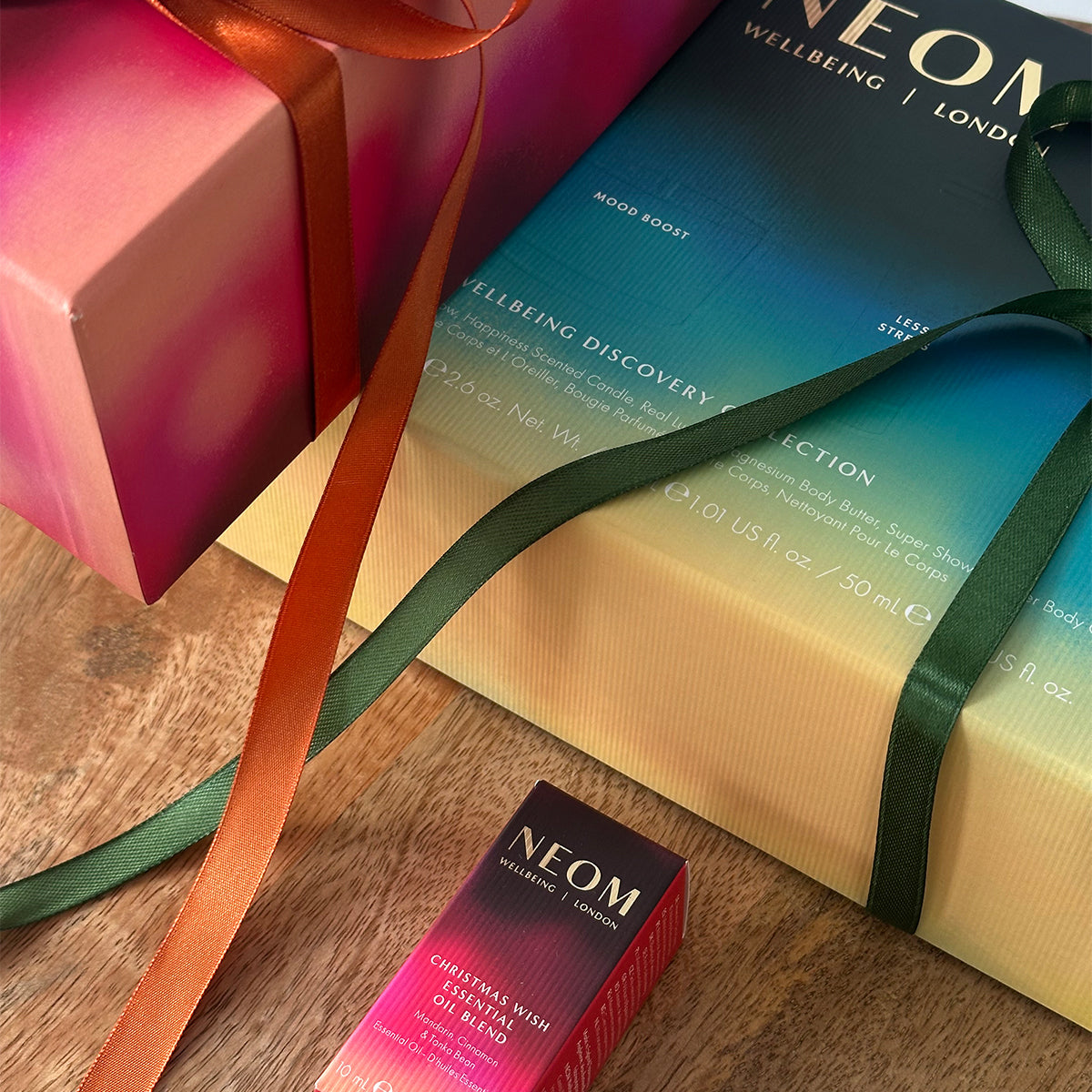 NEOM Christmas gift boxes wrapped with green and orange ribbons beside Christmas Wish Essential Oil Blend.