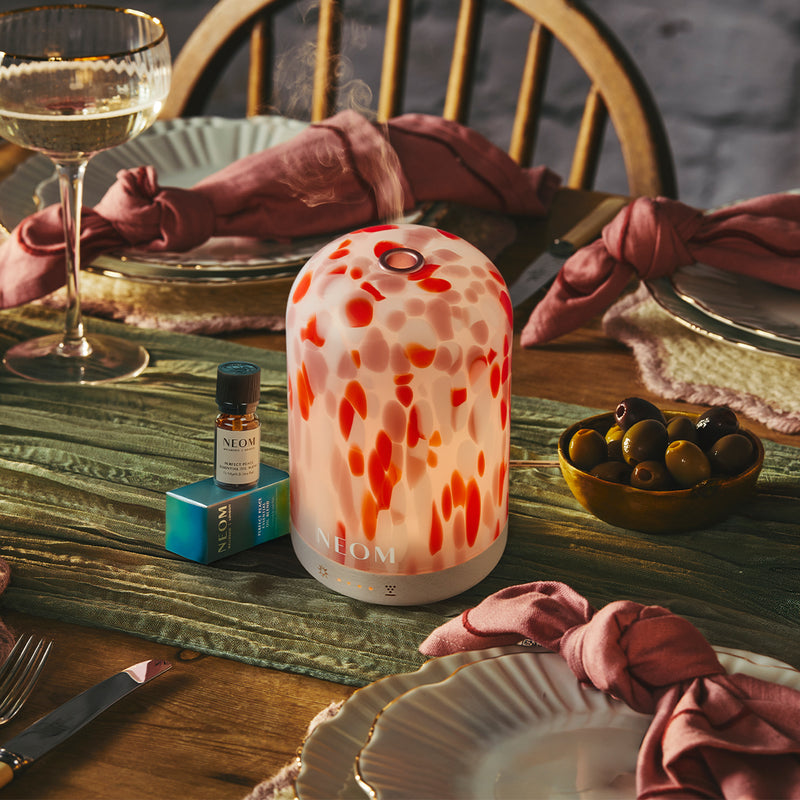 Decorative clementine pod diffuser on a table with wine glasses, olives, and a bottle of essential oil.