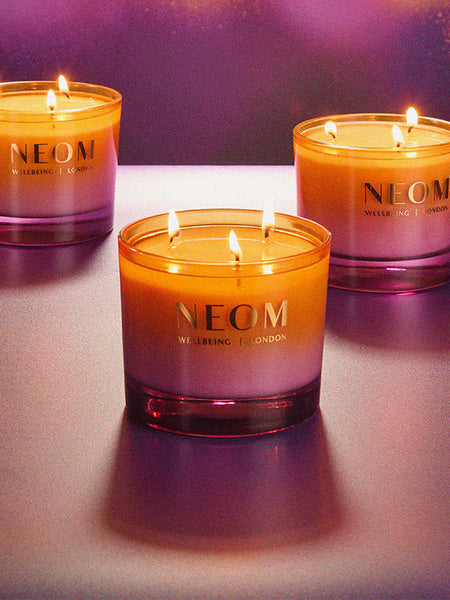 Three NEOM Cosy Nights candles on a reflective surface with a purple gradient background
