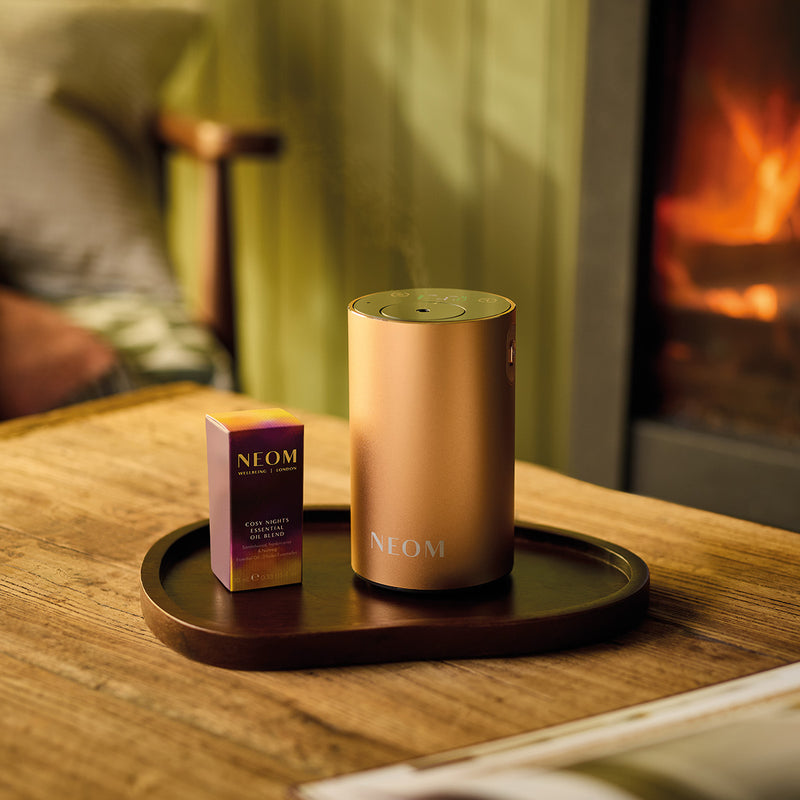 NEOM Rose Gold Pod Mini+ and Cosy Night's essential oil blend wooden table with a fireplace in the background