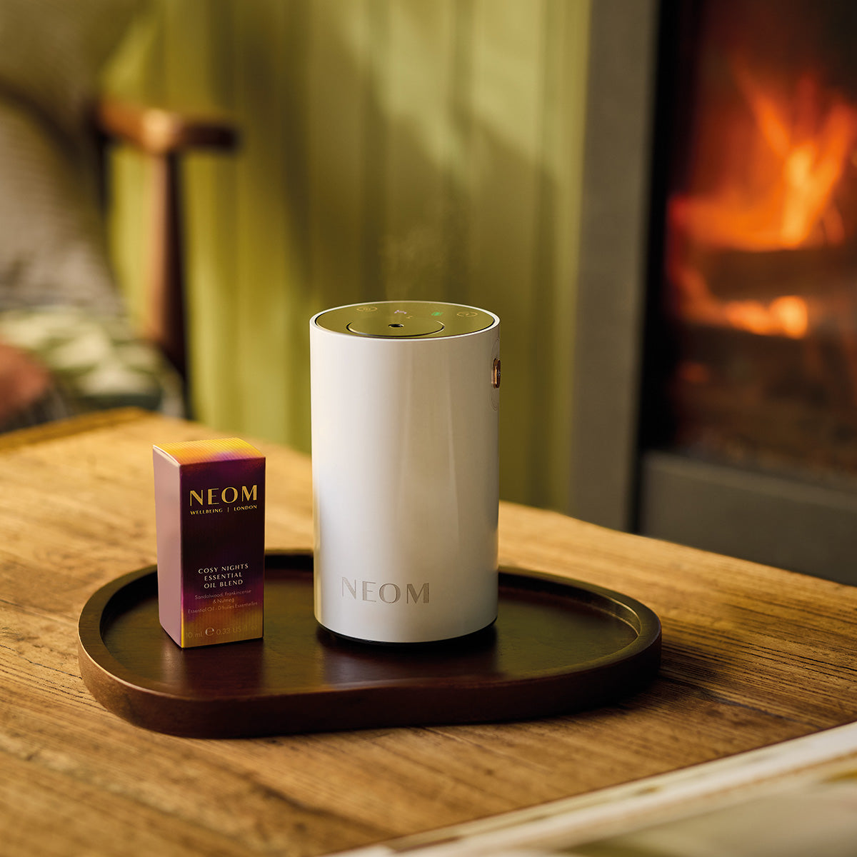 NEOM White Pod Mini+ and Cosy Night's essential oil blend on a wooden tray with a fireplace in the background