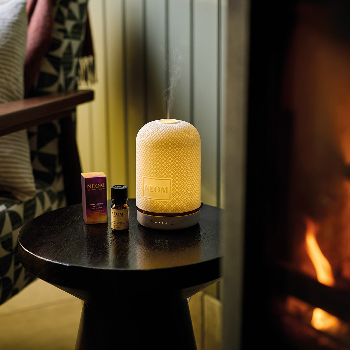 NEOM Wellbeing Pod and COsy Night's essential oil blend on a table with a fireplace in the background