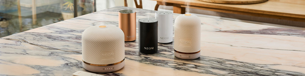 NEOM wellbeing pod & pod luxe essential oil diffusers and Pod Mini+ portable diffusers in black, white and rose gold on marble table.