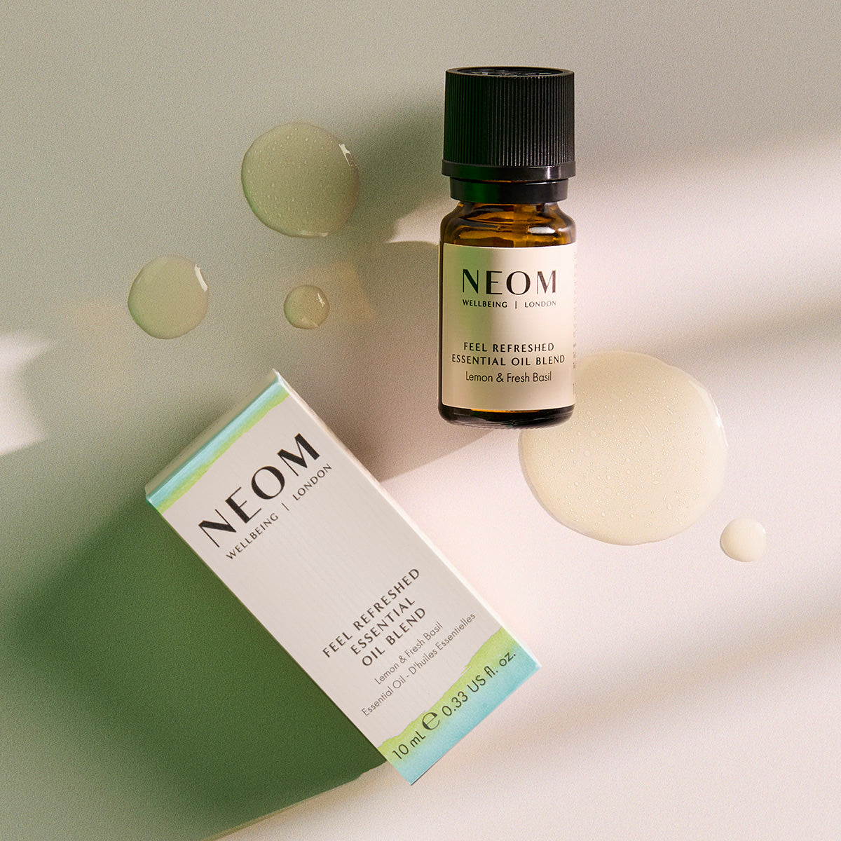 NEOM Feel Refreshed Essential Oil Blend bottle and box placed on a light surface with small pools of oil around them.