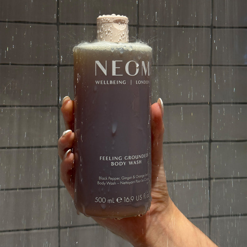Hand holding NEOM Feeling Grounded Body Wash (500 ml) under running shower water against tiled wall