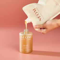 Hand pouring NEOM Great Day hand wash from a large refill bottle into a smaller one against a pink background