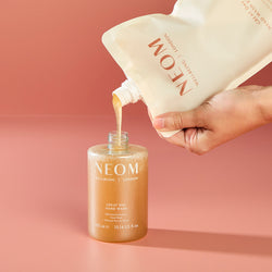 Hand pouring NEOM hand wash into a bottle against a pink background