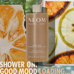 “NEOM Great Day Exfoliating Body Wash (500 ml) centered over a collage of citrus slices and greenery. Text reads: ‘Shower on. Good mood loading…’”