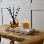 NEOM happiness scented candle and reed diffuser displayed together on a black tray on a rustic wooden bench.
