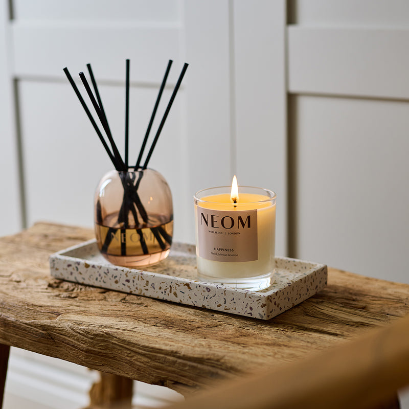 NEOM happiness scented candle and reed diffuser displayed together on a black tray on a rustic wooden bench.