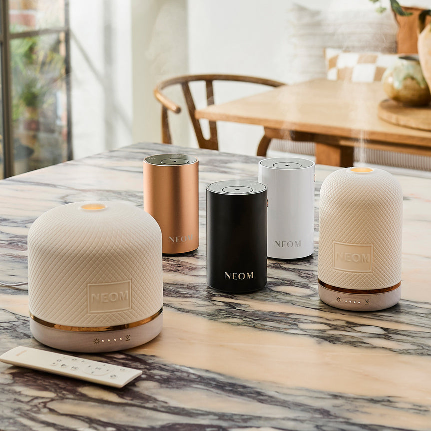 NEOM wellbeing pod & pod luxe essential oil diffusers and Pod Mini+ portable diffusers in black, white and rose gold on marble table.