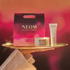 NEOM skincare products including a candle and cream on a tray with a blurred red background