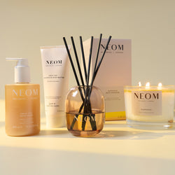 NEOM product range including a bottle, tube, reed diffuser, and candle on a light background
