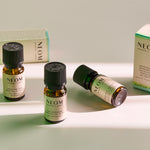 Three NEOM Wellbeing essential oil blend bottles arranged on a light surface with product boxes behind them, softly lit with green-toned shadows.