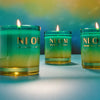 Three lit candles in green glass jars with 'NLCM' branding on a blurred background