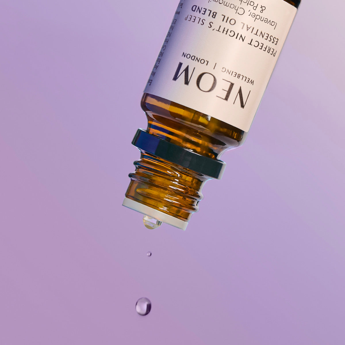 Close-up of an inverted NEOM essential oil bottle releasing drops of oil against a purple background.