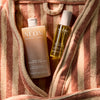 NEOM Great Day Exfoliating Body Wash and Great Day Pro-Vitamin D3 Dry Body Oil inside a striped towel bag.