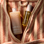 NEOM Great Day Exfoliating Body Wash and Great Day Pro-Vitamin D3 Dry Body Oil inside a striped towel bag.