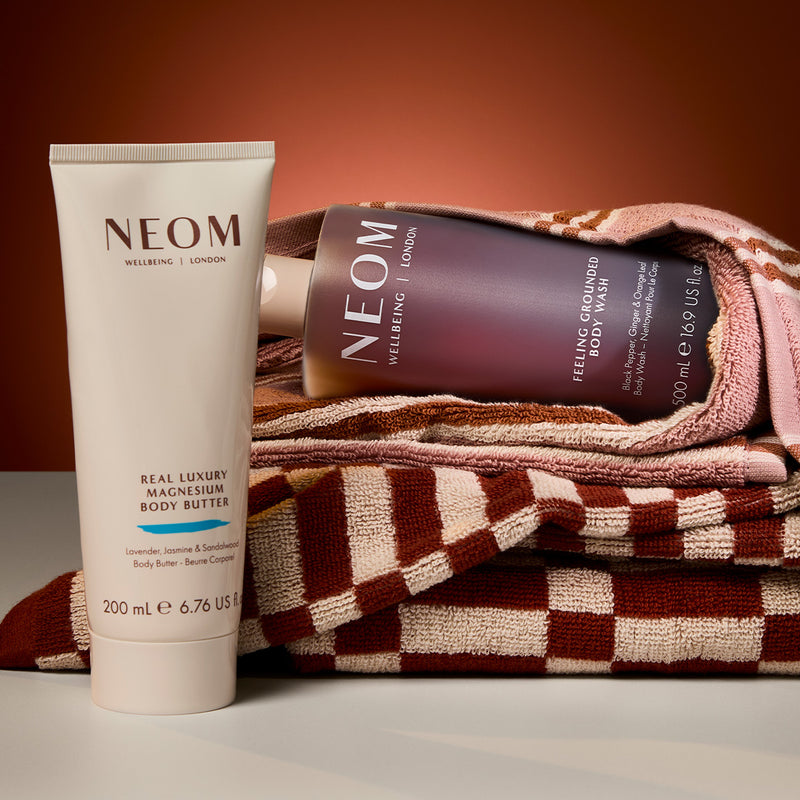 “NEOM Real Luxury Creamy Body Wash and magnesium body butter bottle resting on soft, textured fabric with warm light.