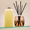 NEOM Squeeze the Day Body Wash next to a NEOM reed diffuser on a glass shelf