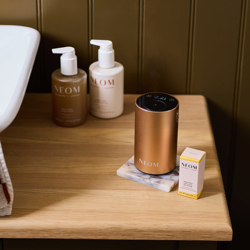 NEOM pod mini+ in rose gold, essential oil blend, hand wash and hand lotion in a a bathroom beside a sink
