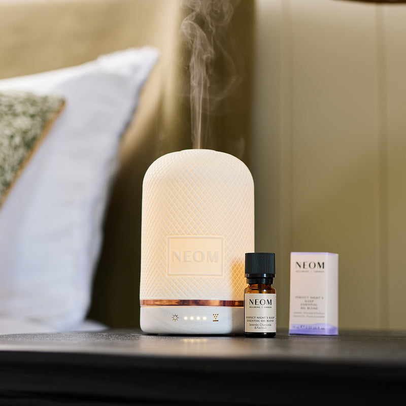 NEOM Wellbeing Pod diffuser releasing mist beside a bottle of Perfect Night’s Sleep essential oil blend on a bedside table.