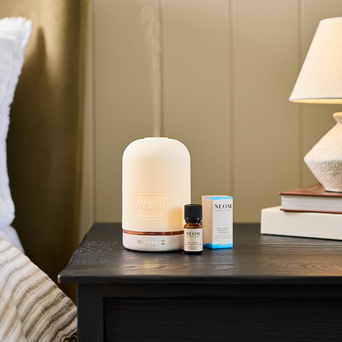 NEOM Wellbeing Pod diffuser releasing mist beside a bottle of Perfect Night’s Sleep essential oil blend on a bedside table.