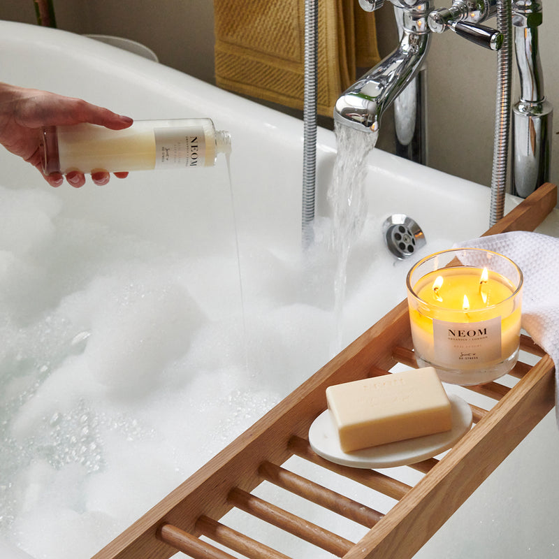 Person holding a NEOM product over a bathtub with a candle and soap on a tray.