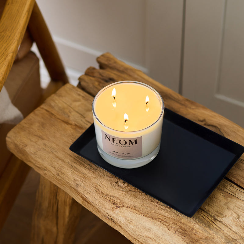 NEOM Real Luxury three-wick scented candle burning on a black tray on a rustic wooden bench.