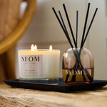 NEOM Real Luxury scented candle and reed diffuser displayed together on a black tray on a rustic wooden bench.