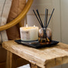 NEOM real luxury scented candle and reed diffuser displayed together on a black tray on a rustic wooden bench.