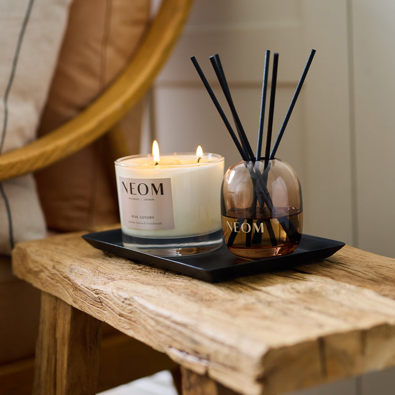 NEOM real luxury scented candle and reed diffuser displayed together on a black tray on a rustic wooden bench.