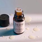 Open NEOM Real Luxury essential oil blend bottle surrounded by small drops of oil on a light surface.