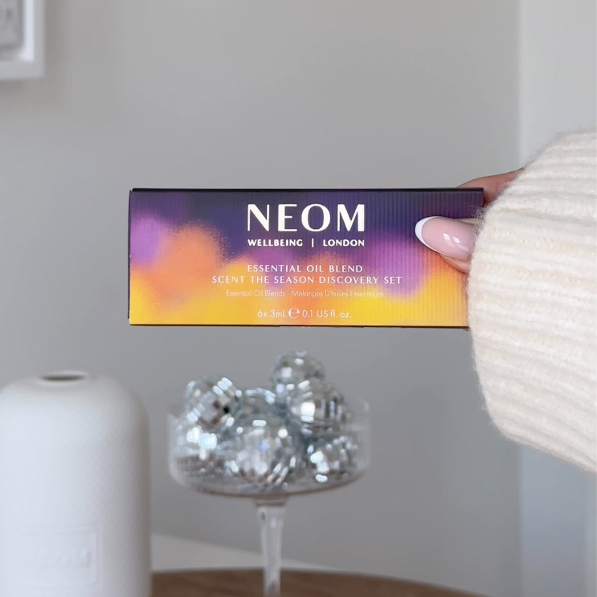 Hand holding NEOM Scent the Season Discovery Set box with essential oil blends and silver disco balls.