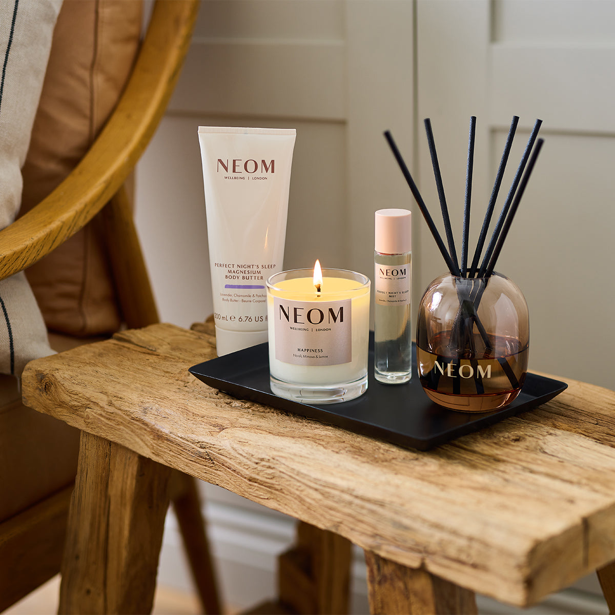 NEOM wellness products including Perfect Night’s Sleep Magnesium Body Butter, Happiness candle, sleep mist and reed diffuser on a wooden bench.
