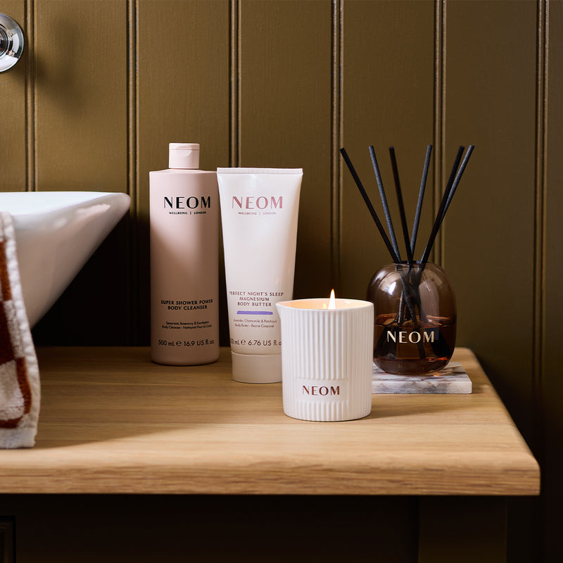 NEOM Super Shower Power Body Cleanser, Magnesium Body Butter, scented candle and reed diffuser styled together on a wooden bathroom shelf.